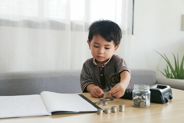 Little boy is saving money, counting coins and putting them in a glass jar. He learned to save money for his education and to buy things he wanted in the future.
