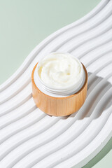Jar with creamy cosmetic product, cream on white tray on green background. Unbranded package with of antioxidant cream for self-care. View from above.