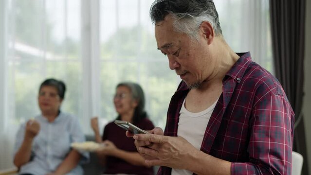 Handheld video of a tech-savvy senior Asian man using his smartphone, oblivious to the two women chatting nearby. The bright living room setting adds to the feeling of connection and enjoyment