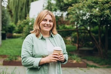 Happy redhead 30s Woman holding brown paper cup of coffee on public park background, Coffee to go on the city street. Food, rest, Take away concept.