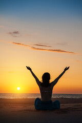 Fototapeta premium Woman exercising yoga while doing meditation at beach during sunrise or sunset sitting on the sand with hands up against sea water and sky