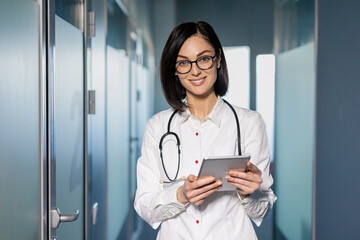 Confident female doctor in white coat holding tablet standing in modern hospital hallway....