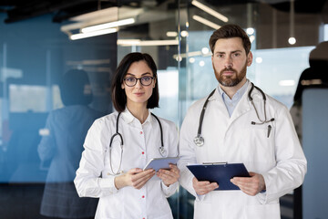 Two doctors in white coats standing in a modern hospital setting. The male doctor holds a clipboard, and the female doctor uses a tablet. Projecting professionalism and medical expertise.