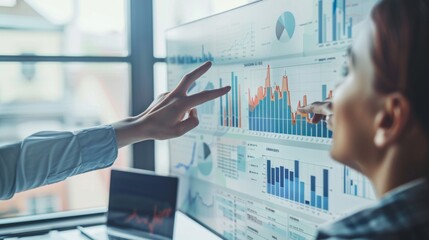Two professionals analyzing business data on a screen, discussing financial graphs and charts in a modern office environment.