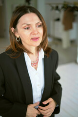 A woman dressed in a black blazer and white blouse carefully adjusts her necklace, showcasing her refined style and professional elegance. Her composed and thoughtful expression adds to her