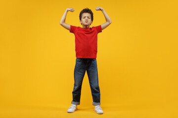 Full length studio portrait of cute confident African American schoolboy posing over bright colored orange yellow background, showing his biceps. Sports, active lifestyle, children health care concept