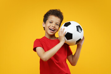 Studio portrait happy smiling beautiful African American boy kid wearing basic red t-shirt, posing with soccer ball in hands, isolated over bright colored orange yellow background