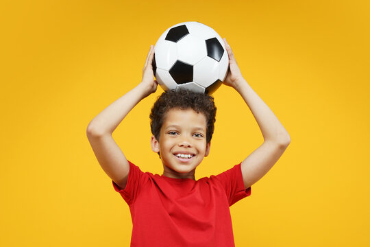 Studio portrait of happy smiling beautiful African American boy kid wearing basic red t-shirt, posing holding soccer ball over his head, isolated over bright colored orange yellow background