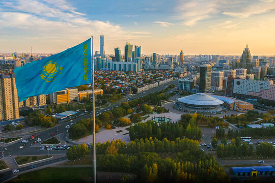 The Kazakhstan flag waiving above Astana city, with the panorama view of the city downtown, the Capital circus, green park, business buildings and skyscrapers, and residential buildings.
