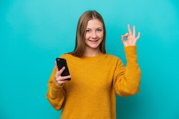 Young Russian woman isolated on blue background using mobile phone and doing OK sign