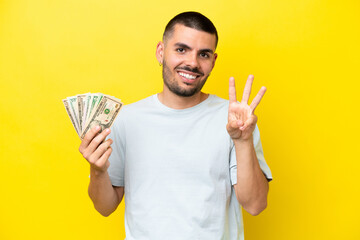 Young caucasian man taking a lot of money isolated on yellow background happy and counting three with fingers