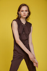 High fashion photo of a beautiful skinny young woman in pretty brown jumpsuit posing on yellow background. Slim figure.