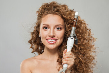 Obraz premium Beautiful young smiling woman holding using hot curling tongs iron on white background. Coiffure, beauty salon and hair styling concept