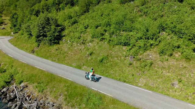 A peaceful women on a ebike ride on a coastal road surrounded by lush forests and clear blue waters on a sunny day. Lovatnet, Norway