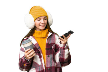 Teenager caucasian girl wearing winter muffs over isolated background holding coffee to take away and a mobile