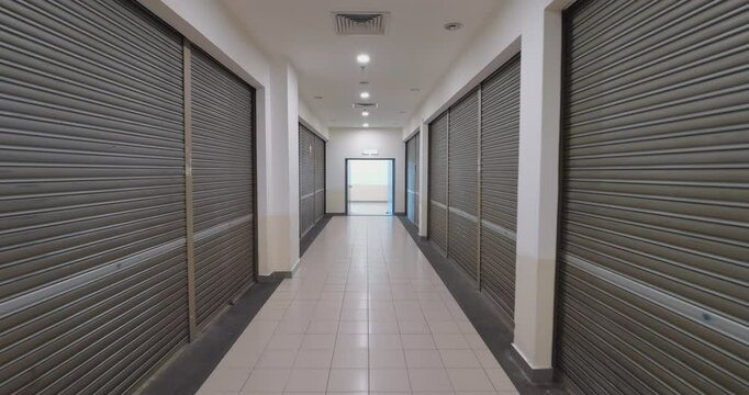 Moving backward through empty corridor with retail spaces closed by rolling shutters, suggesting economic decline and challenges in retail sector. Abandoned shopping center with closed stores