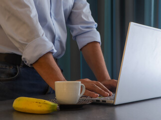 Young businessman typing on his laptop gets up to take a break from work and have a healthy snack consisting of coffee and fruit.