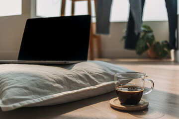 Photo of laptop showing black empty screen, notebook putting on white pillow, on wooden floor in cozy room with black coffee cup. 