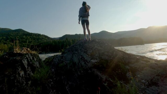 The setting sun illuminates the mountains and the turbulent river. The girl stands on the stone and looks at the river. Dynamic shot with a FPV drone.