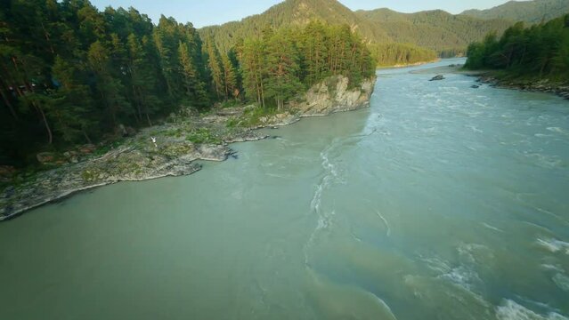 Shooting from a dynamic FPV drone. A flight over a bubbling mountain river with rapids. A flight over the forest and water. Beautiful young woman, standing on a rocky shore.