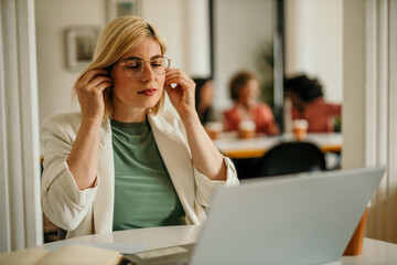 Obraz premium Confident mid aged businesswoman using wireless earphones and looking into a laptop, working in an office