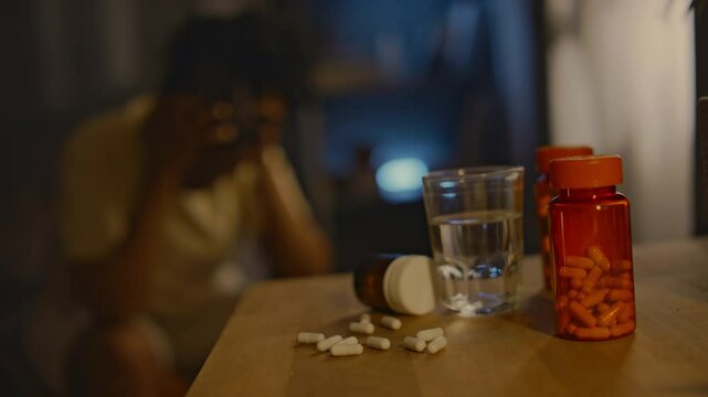 Closeup of plastic jars with sedative pills, glass of water and multiple tranquillizing capsules on wooden table, woman having troubles falling asleep in blurred background, copy space