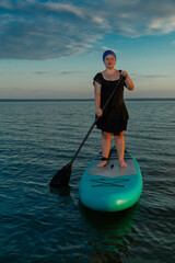 A woman in a closed swimsuit and turban swims on a lake on a paddle board against the backdrop of the sunset sky at dusk