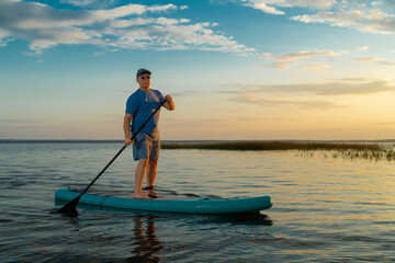 A man in shorts and a T-shirt swims on a lake on a paddle board against the backdrop of a sunset sky with clouds