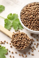 Dried coriander seeds in bowl, spoon and green leaves on light cloth, above view