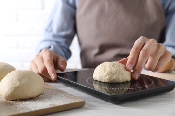 Woman weighing raw dough ball on kitchen scale at white wooden table, closeup