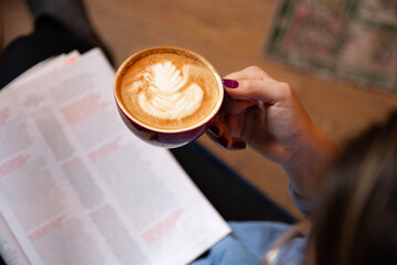 Female hand holding an espresso cup, with a newspaper on her knee. Only the coffee and hand are in focus. Close-up shot