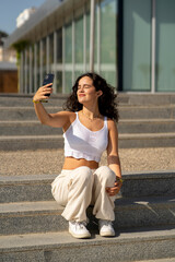 Chica joven en verano sonriendo haciendo fotos con el móvil en un lugar de mar y bonito. Pelo rizado y brillante. 