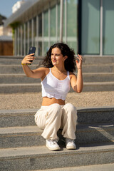Chica joven en verano sonriendo haciendo fotos con el móvil en un lugar de mar y bonito. Pelo rizado y brillante. 