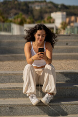 Chica joven en verano sonriendo haciendo fotos con el móvil en un lugar de mar y bonito. Pelo rizado y brillante. 