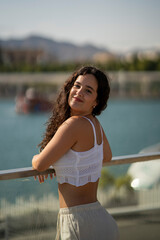 Chica joven en verano sonriendo haciendo fotos con el móvil en un lugar de mar y bonito. Pelo rizado y brillante. 