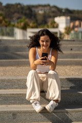 Chica joven en verano sonriendo haciendo fotos con el móvil en un lugar de mar y bonito. Pelo rizado y brillante. 