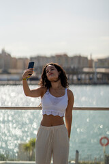Chica joven en verano sonriendo haciendo fotos con el móvil en un lugar de mar y bonito. Pelo rizado y brillante. 