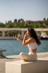 Chica joven en verano sonriendo haciendo fotos con el móvil en un lugar de mar y bonito. Pelo rizado y brillante. 
