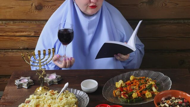 A Jewish woman in a blue veil with glasses of wine and siddur reads kiddush for wine on the Pesach Seder holiday