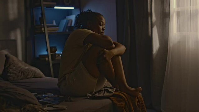 Side full shot of young Black woman with ptsd sitting in bed at night and hugging knees, having compulsive thoughts about past traumatic situations, photos on bed