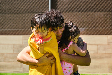 A man is hugging his children, one of whom is eating a watermelon. The man is holding the children close to him, and they are all smiling. Scene is happy and warm