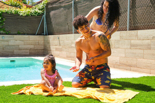 A woman is applying sunscreen to her husband near their daughter next to a pool. The man is wearing shorts. Scene is happy and playful - Powered by Adobe