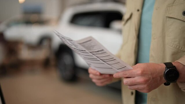 Close-up hands of unrecognizable pensive male customer holding papers, checking vehicle characteristics during choosing car in modern dealership center. Concept of buying new auto at showroom.