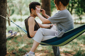 In a serene park setting, a young woman attentively applies makeup to a relaxed man lounging in a...