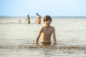 A young boy playfully kicks up water while sitting in the shallow water at the beach, creating a splash. In the background, people enjoy the sunny day.