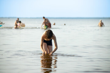 Fototapeta premium A young boy stands in shallow water at the beach, focused on the water in his hands. In the background, several people enjoy the water on a sunny day. Copy space.