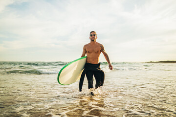 A man wearing a wetsuit and sunglasses walks out of the ocean with a surfboard under his arm. The sun is setting in the background, casting a warm glow on the water.