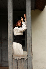 Slender brunette woman in vintage dress and hat standing on an old wooden porch of a school