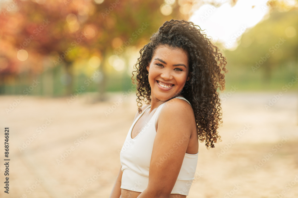 Wall mural A young woman with curly brown hair is smiling and looking off to the side. She is wearing a white tank top and is standing in a park with trees and grass in the background. - Wall murals