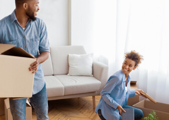 Moving Concept. Joyful African American Spouses Packing Stuff In Boxes Indoor Preparing For Relocation. Selective Focus